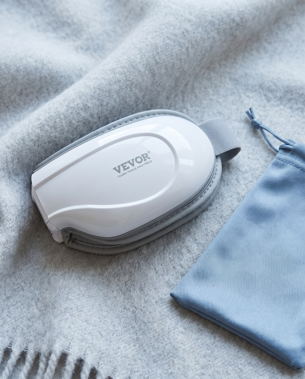 The Heated Eye Massager folded flat on a grey wool throw beside its soft travel pouch.